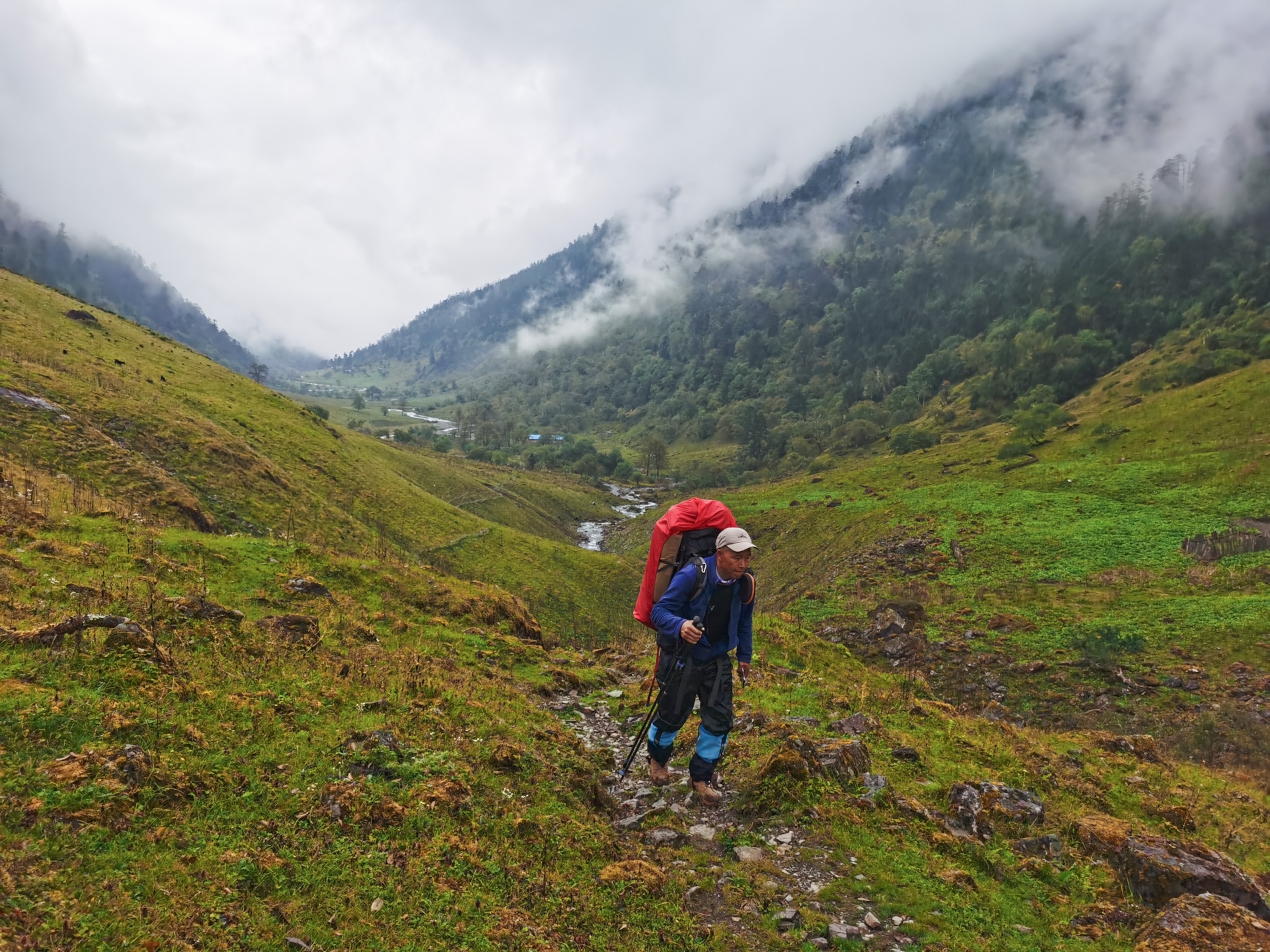 A man wearing a bright red backpack hikes across a lush green trail surrounded by green mountains. Behind him are clouds that cover the top of the mountain peaks.