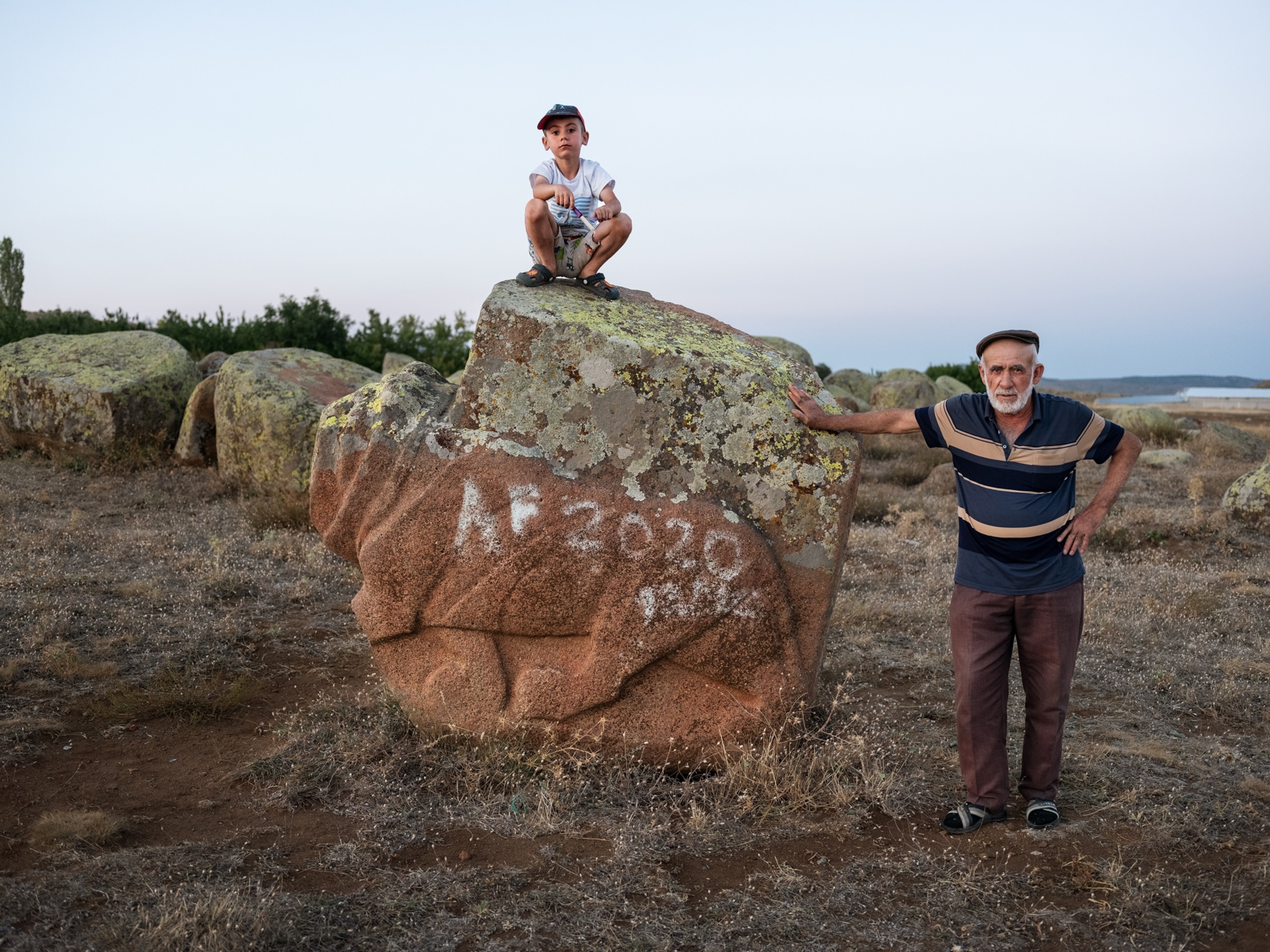 A young boy holding a knife squats on top of an unfinished lion sculpture made of rock. On the right, his grandfather stands leaning against the sculpture.