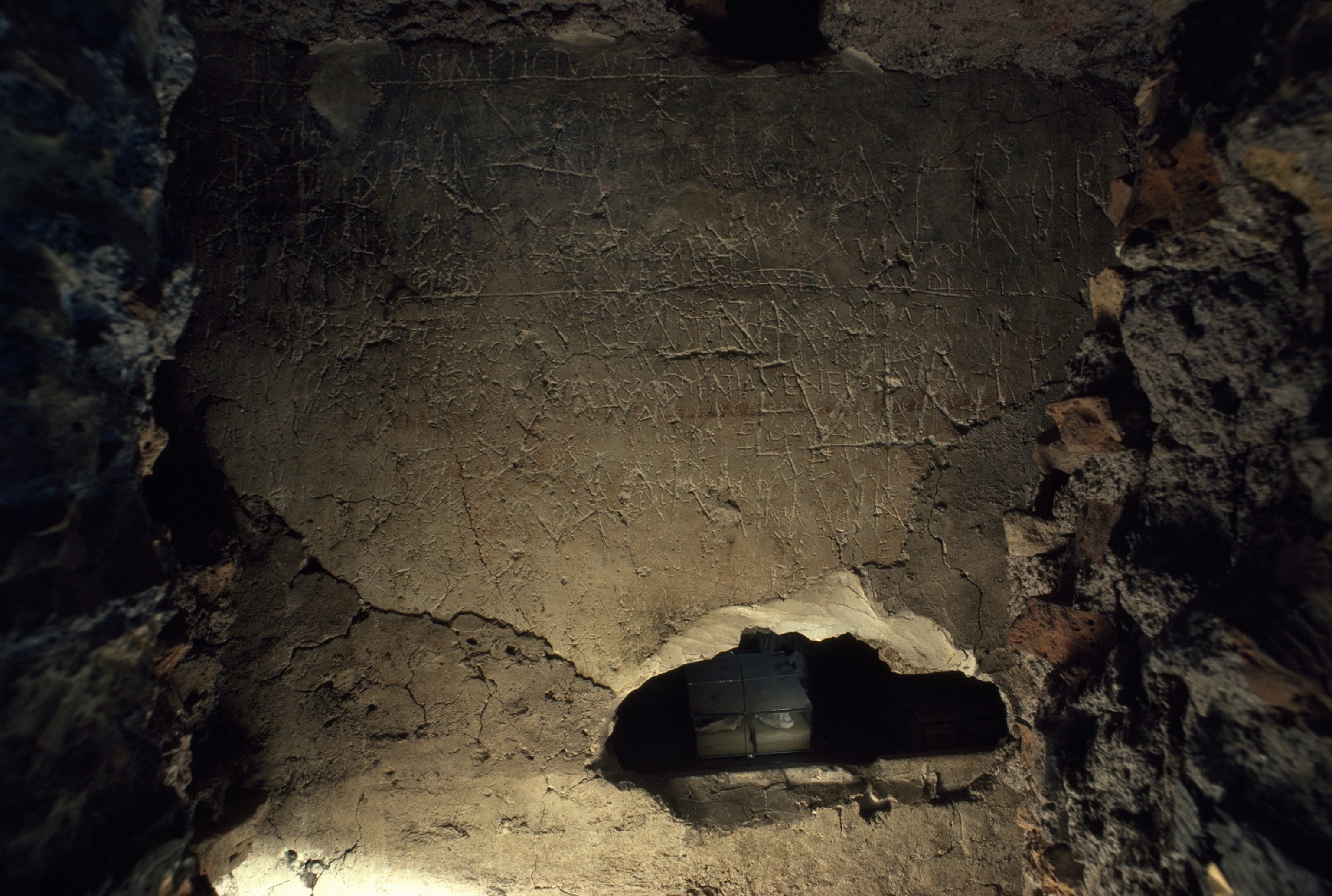 Behind a layer of crumbled brick, a wall shows etchings of words on stone. Behind a hole in the wall, four small containers containing bones can be seen.